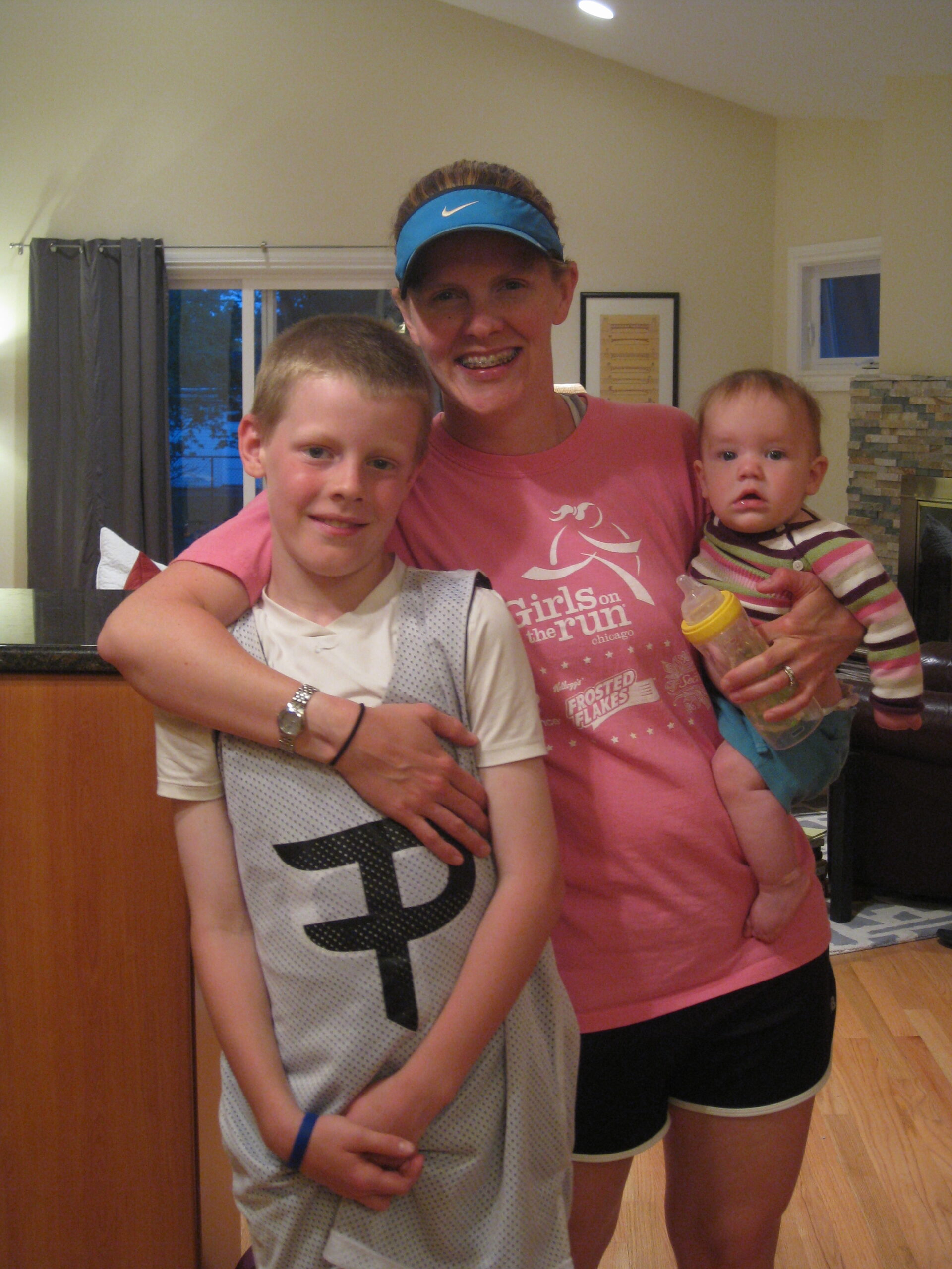 Skinny woman wearing a blue Nike visor, pink T-shirt that says "Girls on the run", and black athletic shorts with white edging, who has her left arm with silver watch and black rubber hair tie on her wrist crossed in front of a young boy who is slightly blocking her while holding in her left arm a toddler.  The young boy has cropped blond hair and is wearing a white T-shirt with an oversized gray athletic vvest with a stylized "P" on it and has a rubber blue bracelet on his left wrist.  The toddler is wearing a brown, white, and pink striped top and blue jeans. In the background is a living room with a white and gray rug, a gray and white brick fireplace, a small window, a piece of artwork on the beige walls, and sliding glass doors with gray curtains.