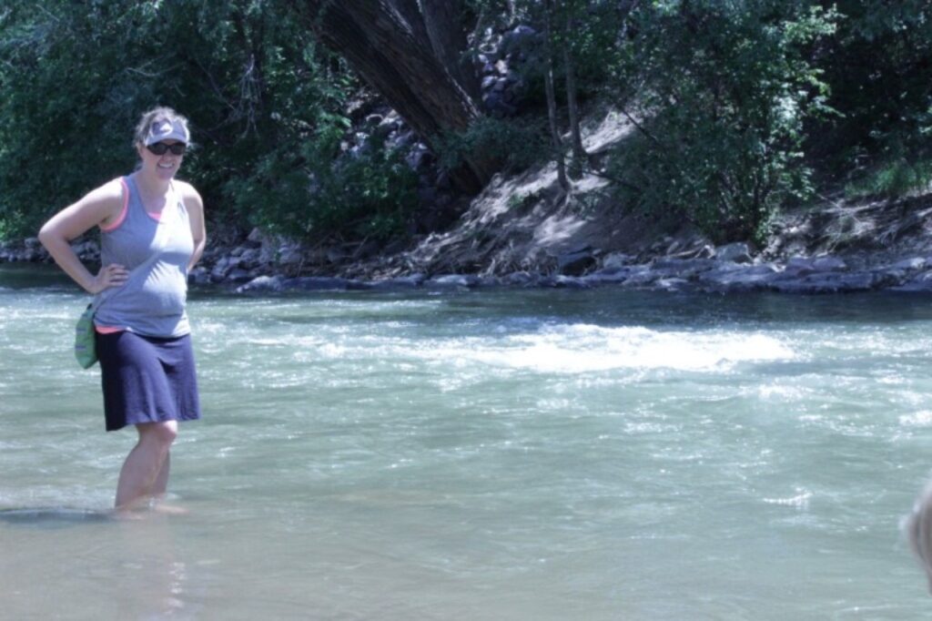 Thin but pregnant woman wearing gray tank top, black visor with white design, dark sunglasses, a navy cotton short skirt, carrying a gray bag on her side, wading in a creek or river with the water up to her shins, with a steep bank and trees on the far side of the water.