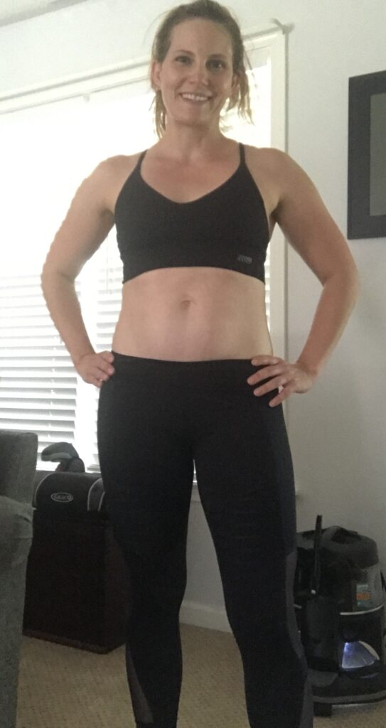 Slender, fit woman named Kelley in black sports bra-like crop top and black spandex work-out pants in front of white wall with window with white blinds pulled closed against sunlight, with two workout  items behind her.