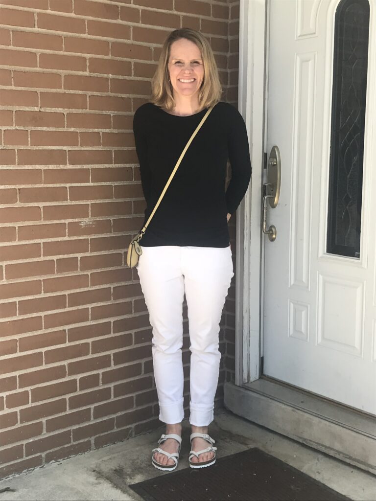 Skinny blonde woman standing on porch in front of white door with brass handle and window with black design and a step between concrete floor and threshold.  She stands in front of a red-brick wall.  She is wearing a black  long-sleeved T-shirt, white skinny jeans, and white leather sandals with straps the cross between her first two toes and has a small tan bag strapped over her chest and hanging on her hip.