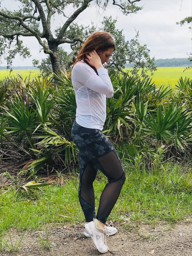 Side full-length shot of woman with brown hair standing facing right in front of tree and shrubberies in spring or summer with a field and hills in the distance and a grayish sky.  She is wearing a transparent white top with a black and white bra-like crop top under it, black and gray athletic leggings mixed with a sheer material on legs, and white athletic shoes with a gray triangle on their sides.
