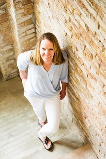 Slender blonde woman shot from above wearing a gray V-necked blouse with 3/4 sleeves, white pants, and brown flip-flops on a wooden floor, leaning against a tan brick wall.